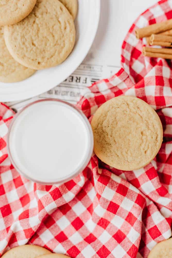 A glass of milk and a sugar cookie sit on a red and white checkered cloth, with more cookies on a white plate and cinnamon sticks nearby.