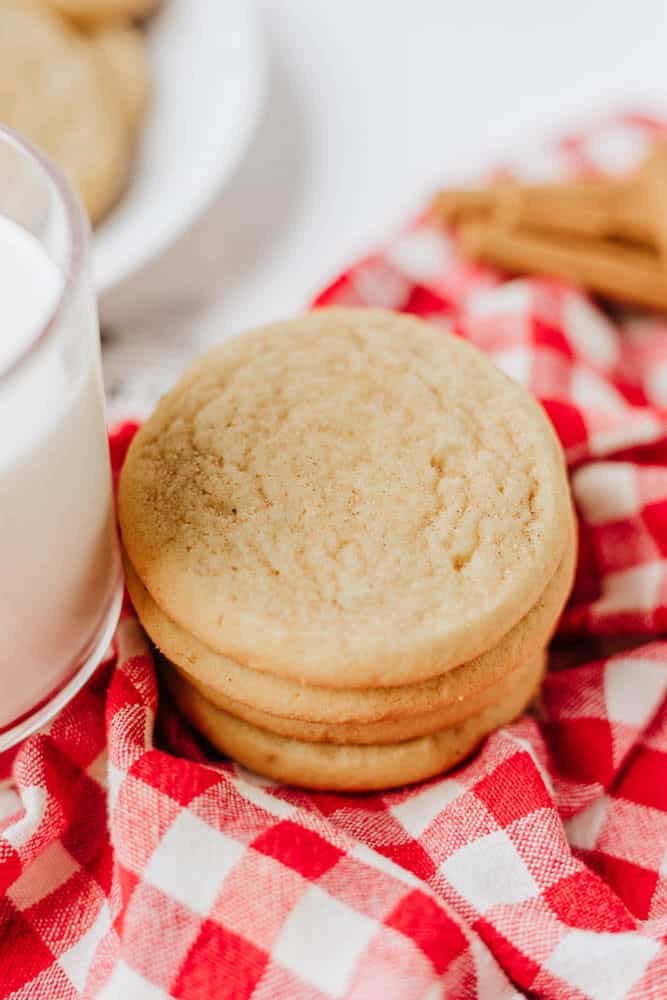 A stack of sugar cookies sits on a red and white checkered cloth next to a glass of milk, with cinnamon sticks and more cookies in the blurred background.