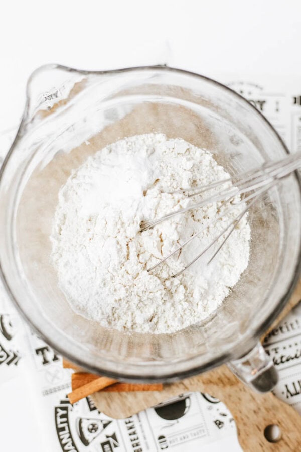 A glass mixing bowl filled with flour and baking powder, with a metal whisk resting inside, sits on a wooden cutting board and newspaper.