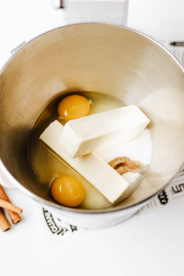 A metal mixing bowl containing two sticks of butter, two raw eggs, brown sugar, and white sugar, ready for baking.