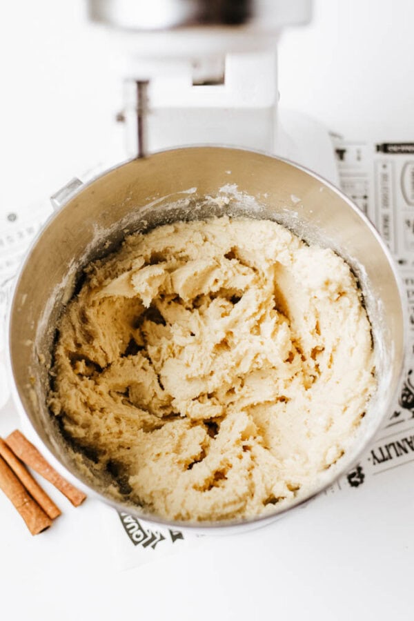 A mixing bowl filled with creamy cookie dough sits in a stand mixer. Two cinnamon sticks and a printed recipe are on the white counter nearby.