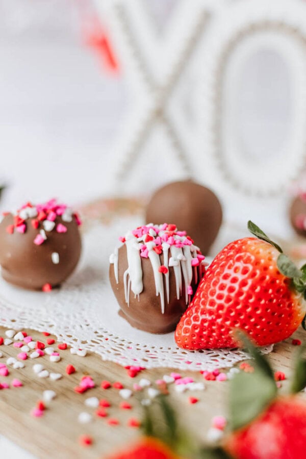Chocolate truffles topped with white drizzle and pink, red, and white sprinkles sit on a doily with a fresh strawberry. A decorative XO sign is blurred in the background.