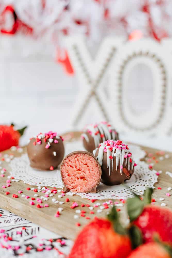 A close-up of four chocolate-covered truffles with pink filling, decorated with sprinkles. One truffle is cut open to show the inside. Strawberries and a decorative XO sign are in the background.