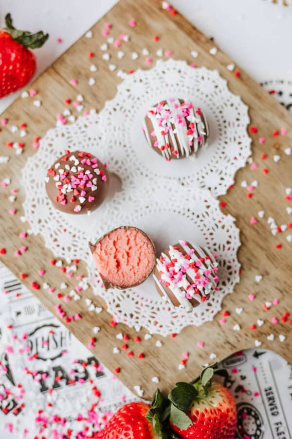 Three decorated cake balls with pink, red, and white sprinkles sit on a wooden board with a doily. One is bitten to reveal a pink center. Fresh strawberries and sprinkles are scattered around.