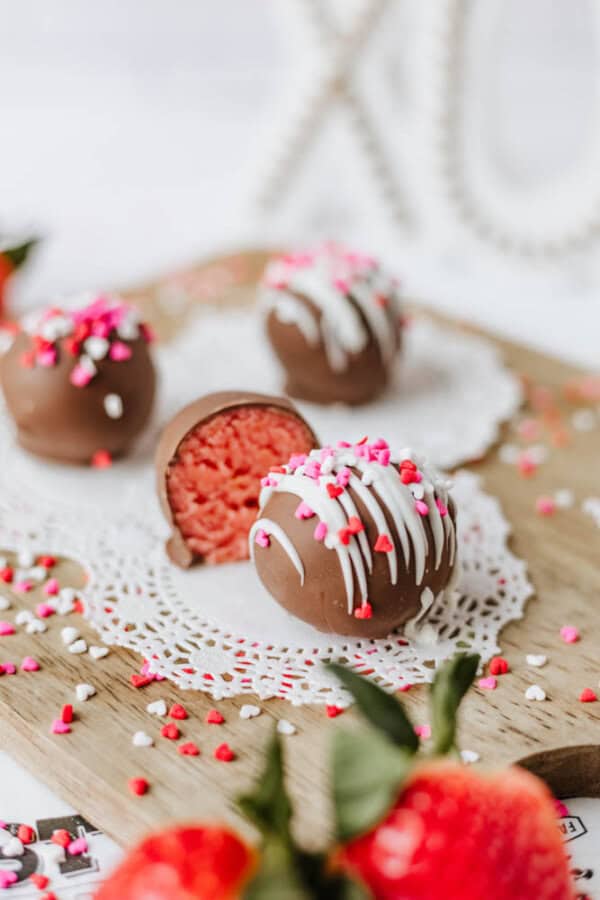 Chocolate-covered cake balls topped with pink, white, and red heart sprinkles are arranged on a wooden board with a lace doily. One cake ball is cut open, showing a pink interior. Fresh strawberries are in the foreground.