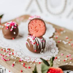 Two chocolate-covered truffles, one with a pink strawberry filling and white drizzle, are stacked on a doily, surrounded by heart-shaped sprinkles and fresh strawberries on a wooden board.