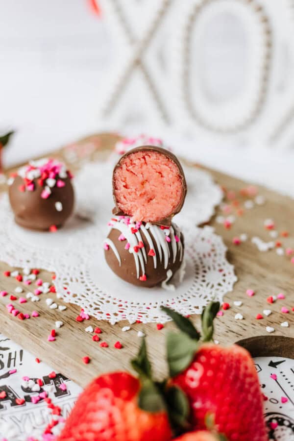 Two chocolate-covered truffles, one with a pink strawberry filling and white drizzle, are stacked on a doily, surrounded by heart-shaped sprinkles and fresh strawberries on a wooden board.