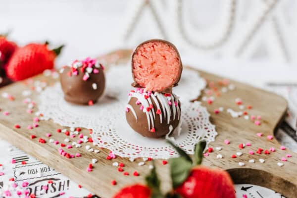 Two chocolate-covered truffles with pink strawberry filling sit on a lacy doily atop a wooden board, surrounded by red, pink, and white heart sprinkles and fresh strawberries.