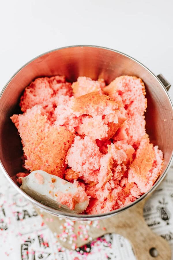 A metal mixing bowl filled with crumbled pink cake pieces, with a spatula partially covered in cake resting inside. The bowl sits on a white surface with a patterned cloth underneath.