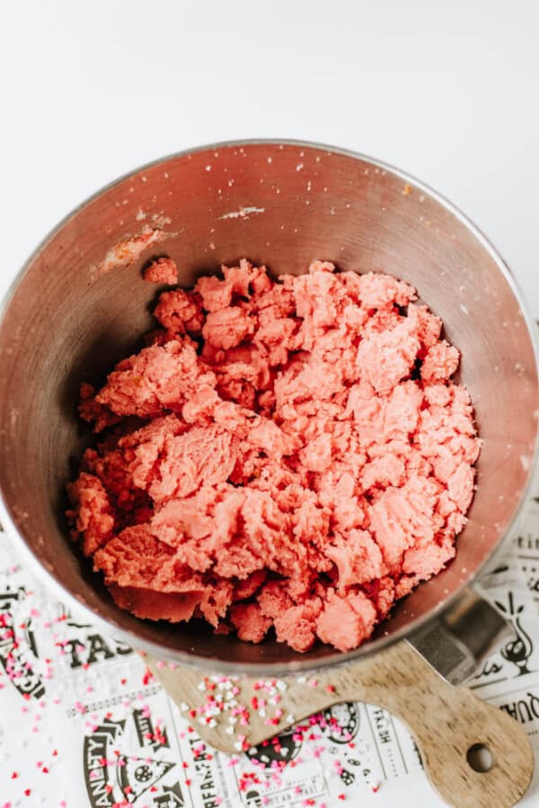 A metal mixing bowl filled with bright pink cookie dough sits on a wooden handle; colorful sprinkles are scattered on a patterned surface nearby.