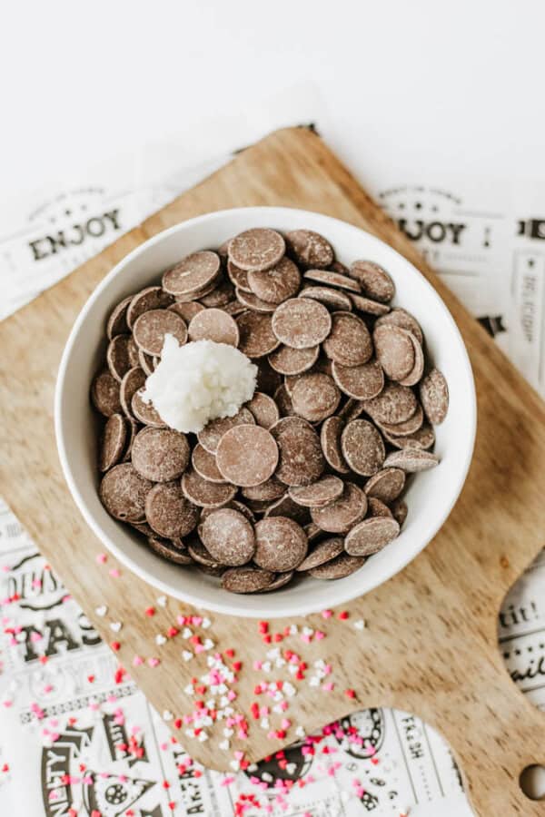 A white bowl filled with chocolate wafers and topped with a single white coconut truffle sits on a wooden board, surrounded by colorful heart-shaped sprinkles and decorative paper.