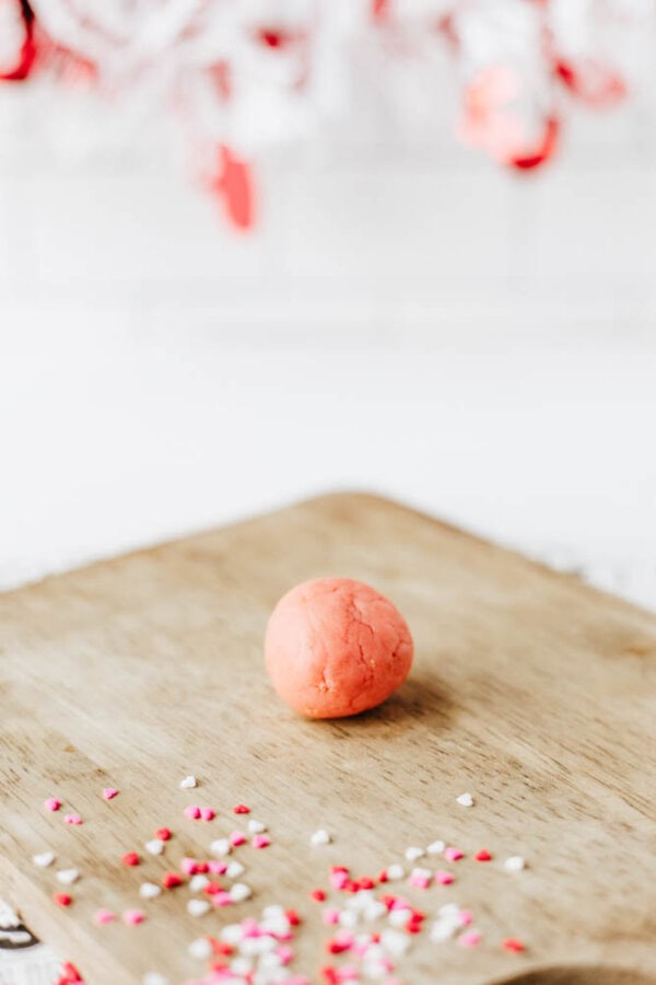 A pink dough ball sits on a wooden surface with small red, pink, and white heart-shaped sprinkles scattered nearby. The background is softly blurred with hints of red and white.