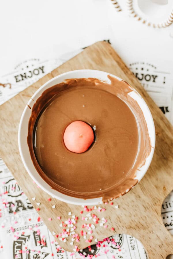 A bowl of melted chocolate with a pink candy sphere in the center, placed on a wooden board. Colorful sprinkles are scattered nearby on a decorative paper.