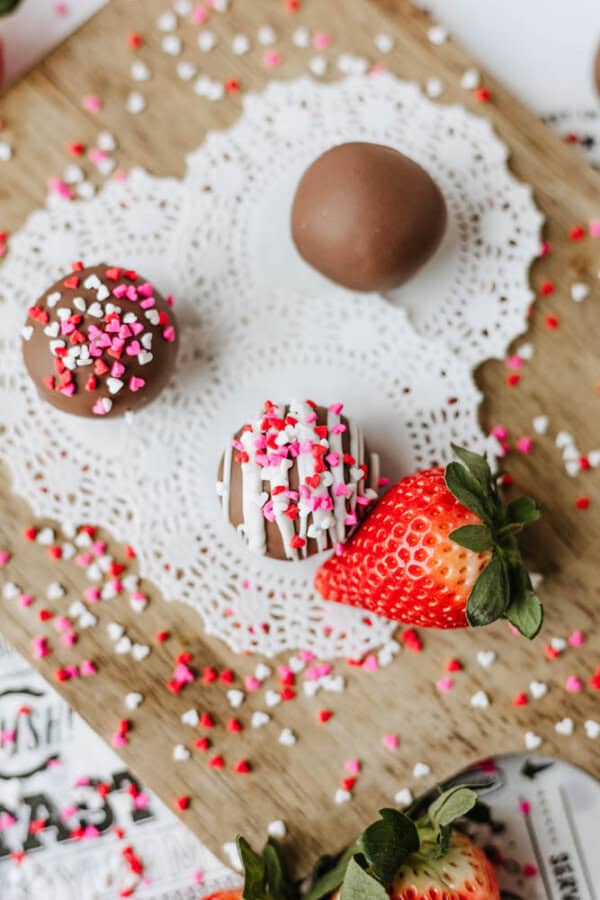 Three chocolate-covered treats, two topped with pink, red, and white heart-shaped sprinkles, sit on a white doily on a wooden board alongside a fresh strawberry. Colorful sprinkles are scattered around.