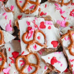 White chocolate bark topped with pretzels, heart-shaped sprinkles, pink and red confetti sprinkles, and white sugar pearls, broken into pieces and arranged closely together.