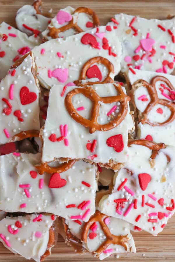 A pile of white chocolate bark topped with pretzels, heart-shaped sprinkles, red and pink sprinkles, and white candy pearls, displayed on a wooden surface.