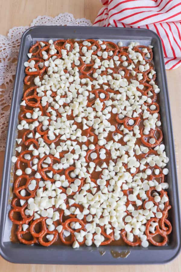 A baking tray filled with pretzels topped with melted caramel and sprinkled generously with white chocolate chips, sitting on a wooden surface with a red-striped towel and lace doily nearby.