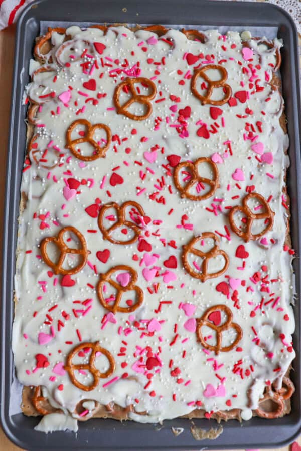 A baking tray filled with a dessert topped with white chocolate, pretzels, pink and red heart-shaped sprinkles, and red and white nonpareils, creating a festive Valentineโs Day treat.