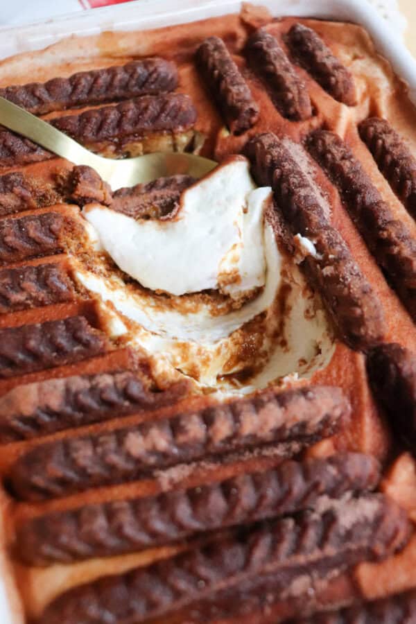 A close-up of tiramisu in a dish, topped with cocoa-dusted ladyfinger biscuits. A spoon has scooped out a portion, revealing the creamy layers underneath.