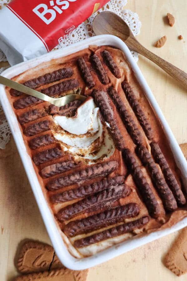 A rectangular dish of layered dessert topped with rows of chocolate cookies, partially scooped out with a gold spoon. A wooden spoon, biscuit package, and loose cookies are next to the dish on a wooden surface.