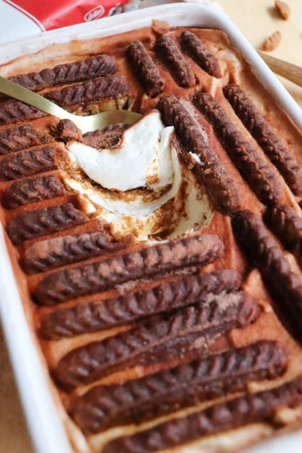 A rectangular dish of tiramisu topped with rows of cocoa-dusted ladyfingers. A spoon has scooped out a portion, revealing creamy layers beneath the surface.