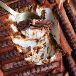 A close-up of a creamy dessert with layers of chocolate biscuits and whipped filling, topped with dusted cocoa powder. A gold spoon has scooped out a portion, revealing the creamy layers inside.