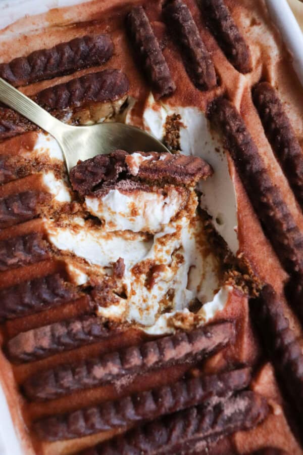 A close-up of a creamy dessert with layers of chocolate biscuits and whipped filling, topped with dusted cocoa powder. A gold spoon has scooped out a portion, revealing the creamy layers inside.