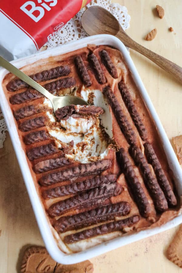 A rectangular dish of dessert, partially eaten, with layers of cream and chocolate cookies arranged on top. A gold spoon is resting in the dish. A package of biscuits is visible in the background.