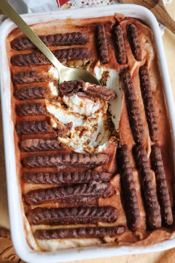 A rectangular dish filled with layered dessert, featuring rows of chocolate biscuits on top and a creamy filling underneath. A spoon has scooped out a portion, revealing the desserts layers.