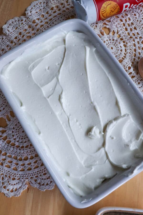A rectangular white dish filled with smooth, creamy white frosting sits on a lacy doily atop a wooden surface. A can of biscuit dough and a wooden spoon are partially visible nearby.