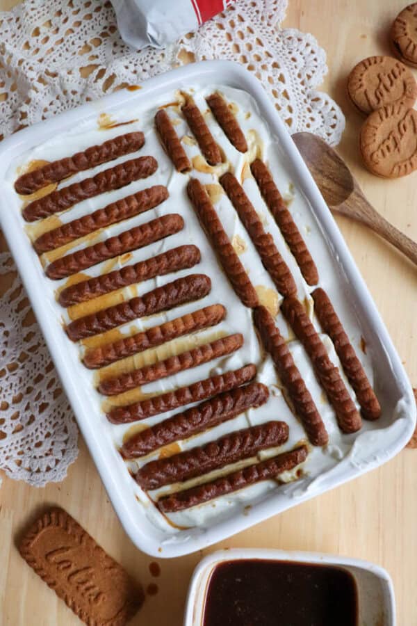 A rectangular dish with a dessert made of layers of cream and neatly arranged rectangular cookies, some slightly soaked. The dish sits on a wooden surface with a lace doily, wooden spoon, and extra cookies nearby.