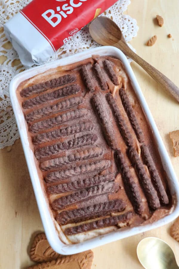 A rectangular dish of chocolate and cream dessert topped with rows of chocolate biscuits. A wooden spoon, cookie pieces, and an opened Biscuit packet are nearby on a light wooden surface.
