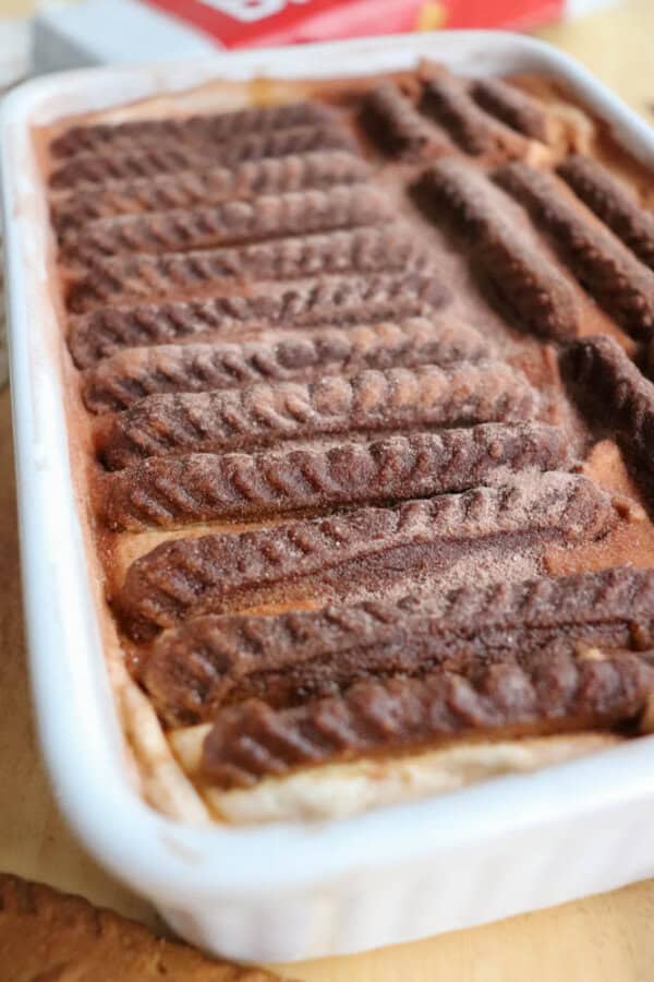 A close-up of a rectangular dessert in a white dish, topped with rows of chocolate cookies or biscuits, lightly dusted with cocoa powder. The dessert has a creamy layer underneath the cookies.