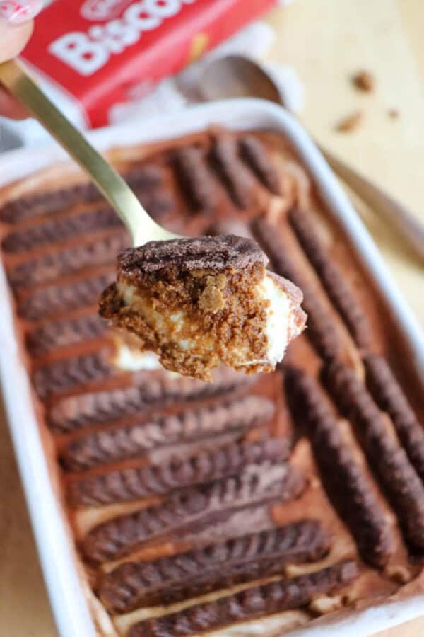 A close-up of a spoon holding a bite of creamy tiramisu with chocolate biscuit topping, held above a baking dish filled with more tiramisu and chocolate biscuits arranged in rows.