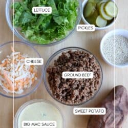 Six labeled ingredients are arranged on a table: a bowl of chopped lettuce, a bowl of sliced pickles, shredded cheese, cooked ground beef, a whole sweet potato, a bowl of Big Mac sauce, and a small bowl of sesame seeds.