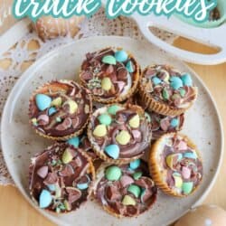 A plate of chocolate-topped cookies decorated with colorful mini candy eggs, sitting on a wooden table. Text on the image reads Mini Egg Crack Cookies So Good! homemadeheather.com.