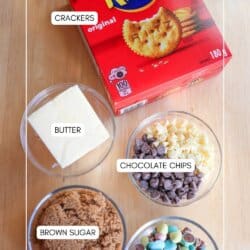 A box of Ritz crackers, a dish of butter, bowls of chocolate chips, brown sugar, and mini eggs arranged on a wooden surface, each labeled with its name.