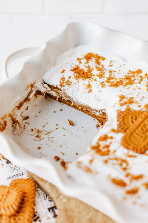 A rectangular white baking dish with a layered biscoff cookie butter delight topped with whipped cream and crumbled cookies, one serving removed to reveal cookie and creamy layers inside. Whole cookies and crumbs are scattered on top and nearby.