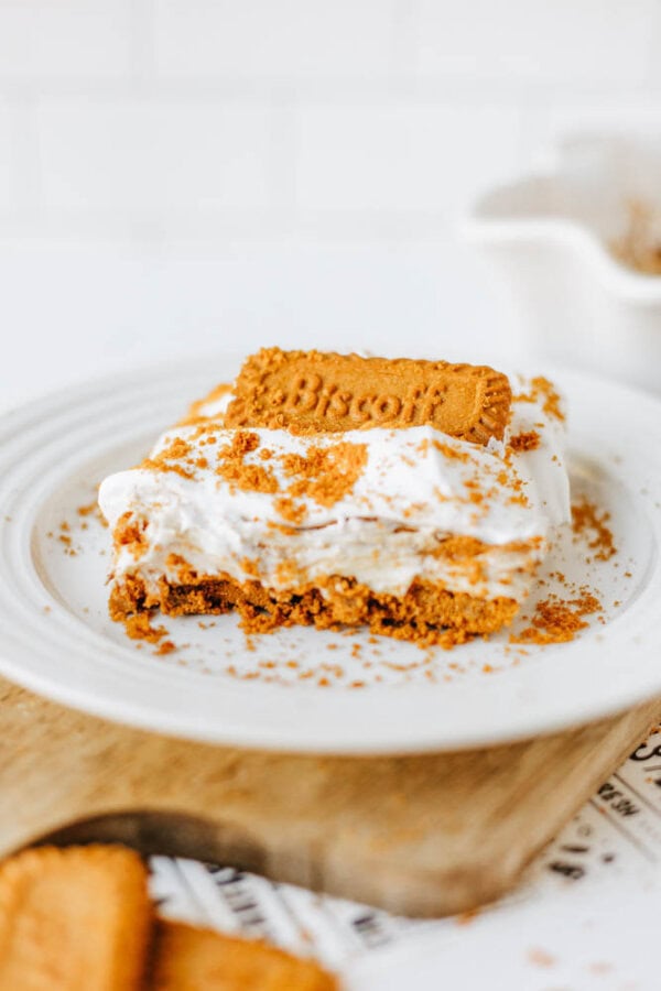 A slice of creamy biscoff cookie butter delight topped with whipped cream, crumbled Biscoff cookies, and a whole Biscoff cookie on a white plate, with more cookies and a baking dish in the background.