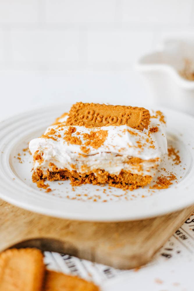 A slice of creamy biscoff cookie butter delight topped with whipped cream, crumbled Biscoff cookies, and a whole Biscoff cookie on a white plate, with more cookies and a baking dish in the background.