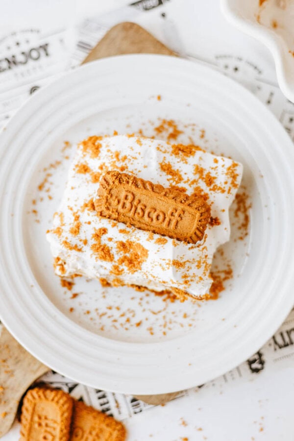 A square piece of creamy Biscoff cookie butter delight topped with whipped cream and Biscoff cookie crumbs, garnished with a whole Biscoff cookie, served on a white plate.