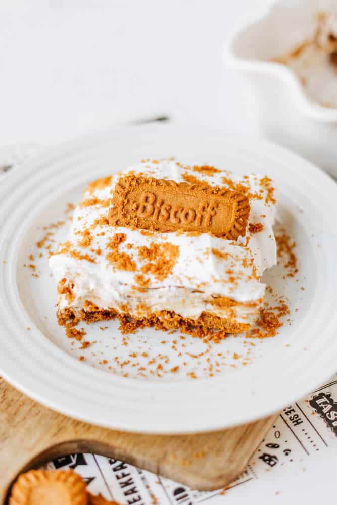 A slice of creamy biscoff cookie butter delight topped with whipped cream and crumbled Biscoff cookies sits on a white plate, garnished with a whole Biscoff cookie on top.