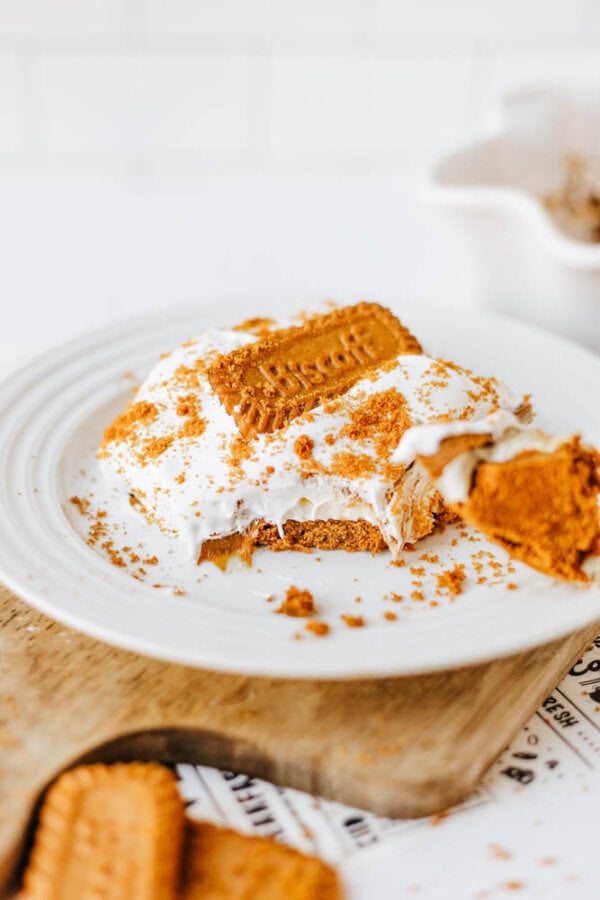 A slice of creamy biscoff cookie butter delight topped with whipped cream and crumbled cookies, garnished with a whole cookie, served on a white plate with a wooden board underneath.