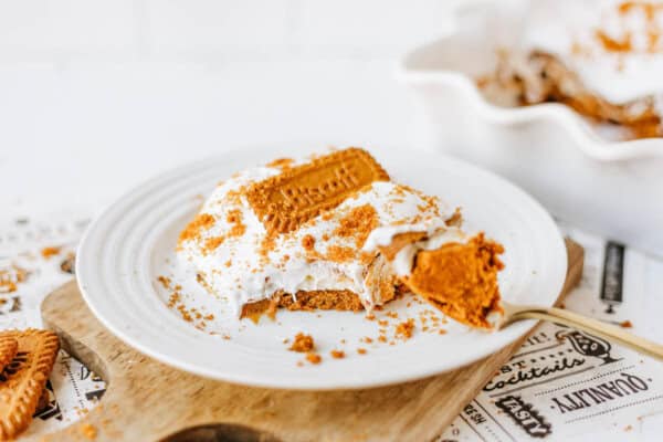 A white plate holds a slice of creamy biscoff cookie butter delight topped with whipped cream, cookie crumbs, and a whole Biscoff cookie. A fork rests beside the dessert on a wooden board, with more dessert in a dish in the background.