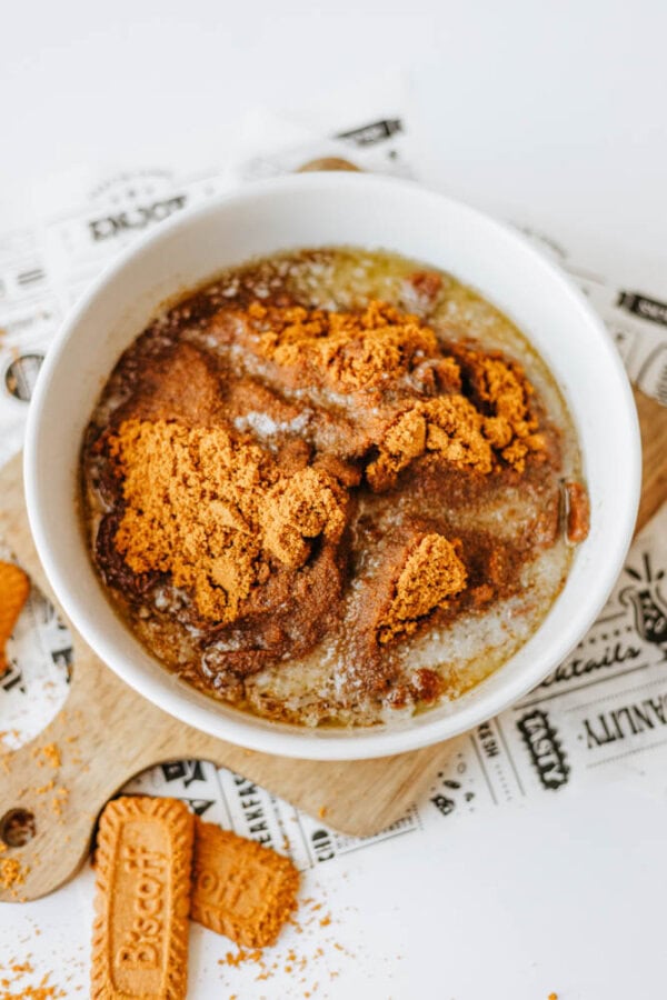 A white bowl filled with oatmeal, topped with crumbled Biscoff cookies, cinnamon, and a drizzle of syrup, sits on a printed napkin. Whole and broken Biscoff cookies add to this biscoff cookie butter delight.