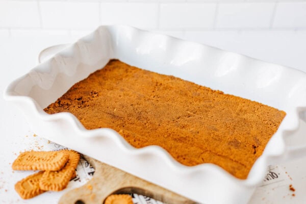 A white rectangular baking dish with a golden brown biscoff cookie butter delight crust evenly spread on the bottom, sitting on a wooden board. Crumbled cookies are scattered nearby.