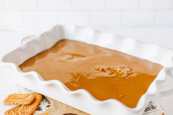 A white baking dish filled with smooth, caramel-colored biscoff cookie butter delight batter sits on a white surface. Nearby, a few rectangular butter cookies are scattered, and the background features a white tiled wall.