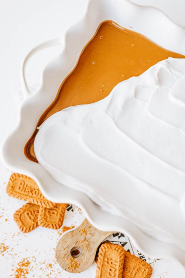 A baking dish filled with a layer of caramel-colored biscoff cookie butter delight, topped with thick whipped cream. Crushed and whole Biscoff cookies are scattered around the dish on a white surface.