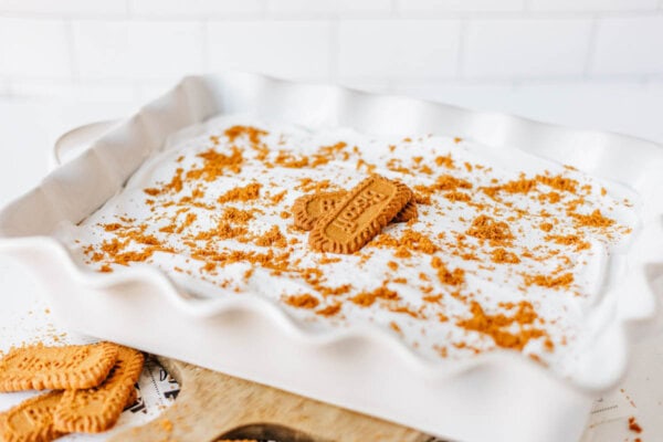 A white ceramic dish filled with creamy biscoff cookie butter delight, topped with crumbled cookies and a whole rectangular cookie in the center, set on a white surface with extra cookies nearby.