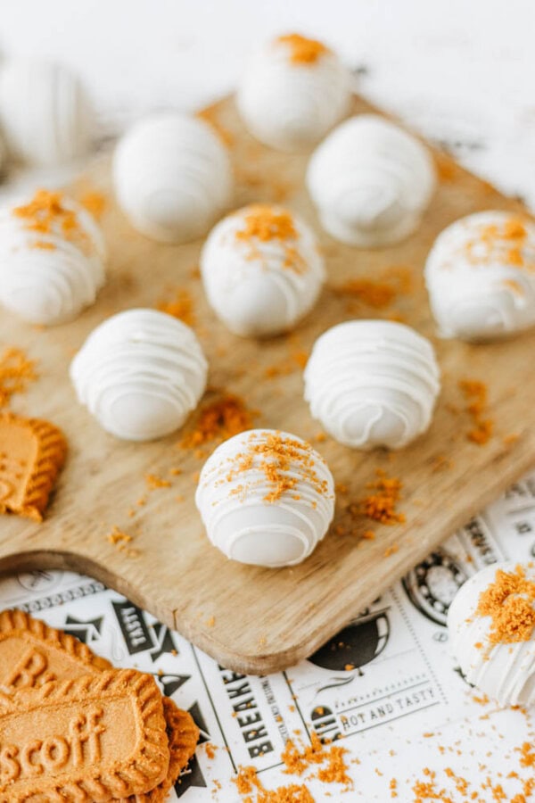 White chocolate-covered truffles arranged on a wooden board, topped with cookie crumbs. Biscoff cookies and crumbs are scattered around, making this a true biscoff cookie butter delight, with a few truffles placed on paper in the background.